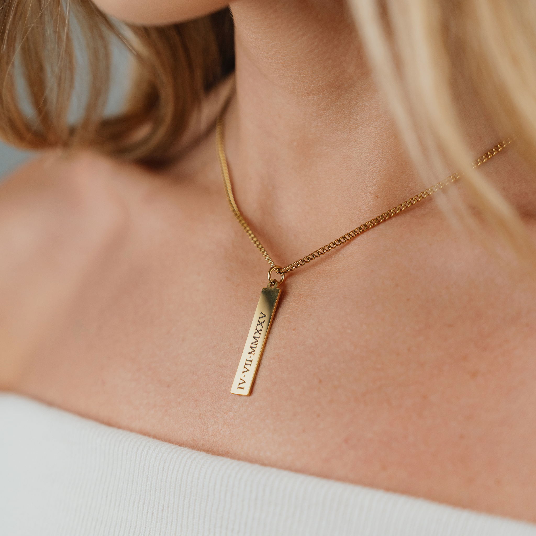 A Female wearing SAAR CO's MEGAN, a polished, 18K Gold plated pendant tag and chain. The tag has been engraved with IV.VII.MMXXV