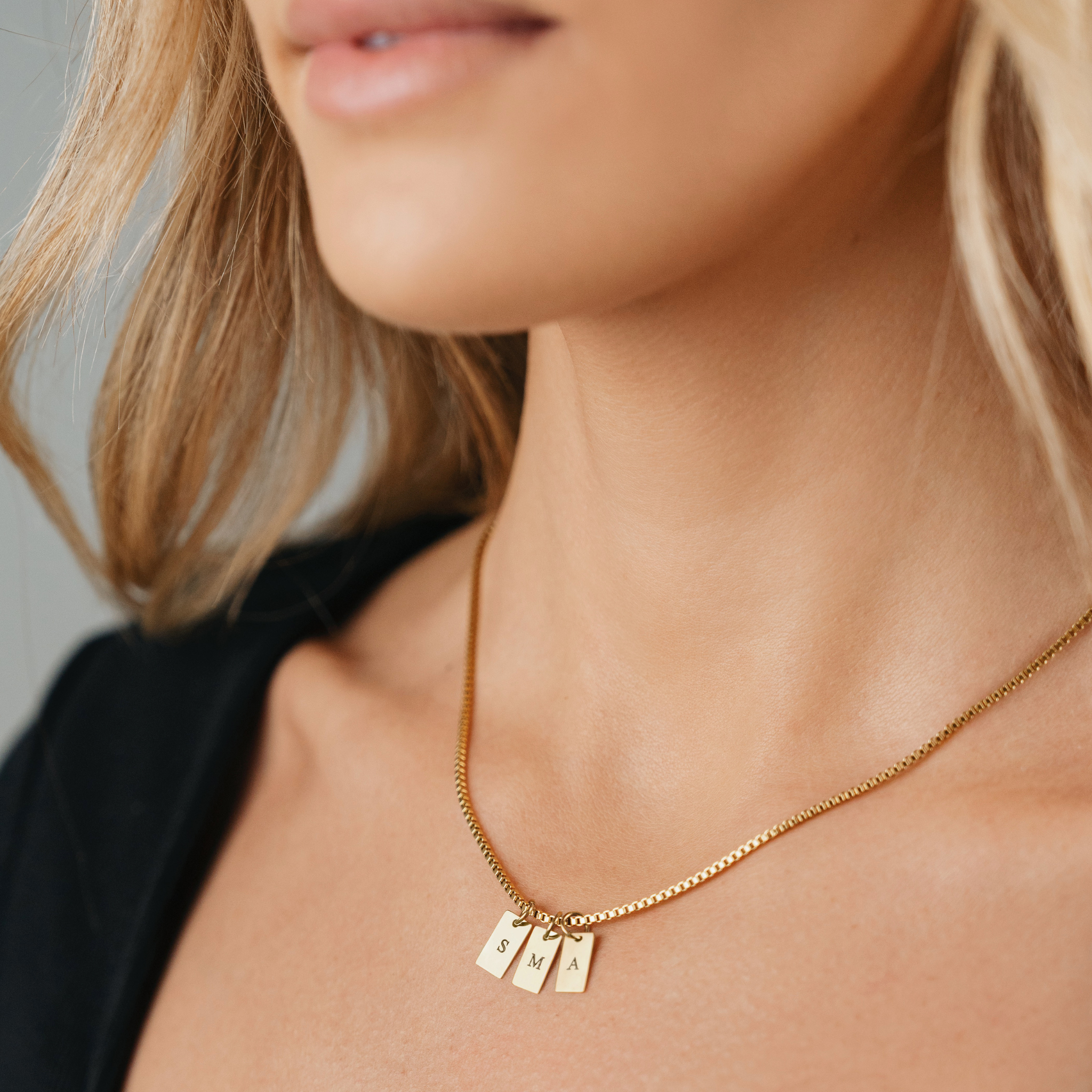 A Female wearing SAAR CO's Elle 18k Gold plaited Stainless Steel pendant chain. The pendant chain includes three pendant tags, which have been engraved with one letter on each tag, the letters are S, M, and A.