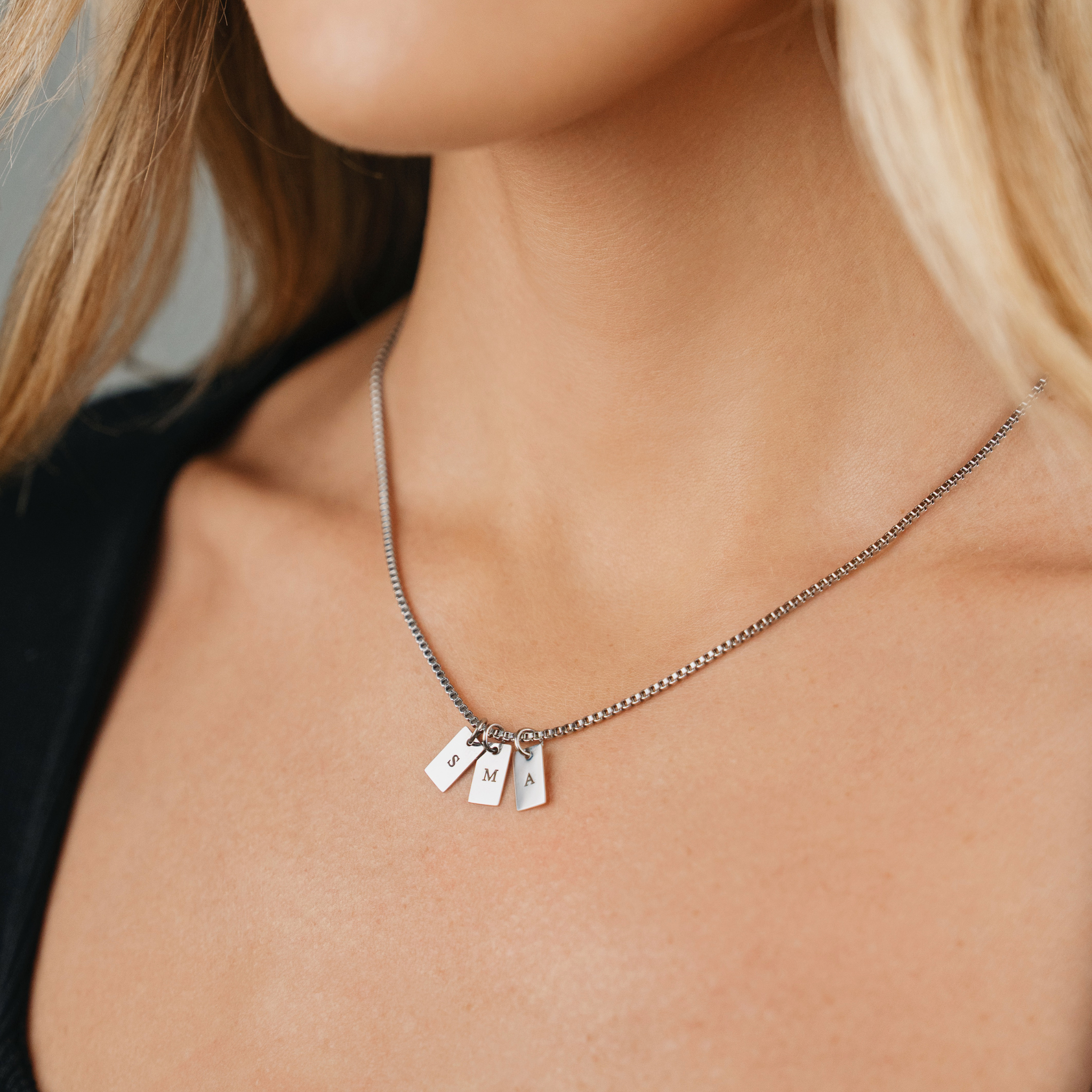 A Female wearing SAAR CO's Elle Stainless Steel pendant chain. The pendant chain includes three pendant tags, which have been engraved with one letter on each tag, the letters are S, M, and A.