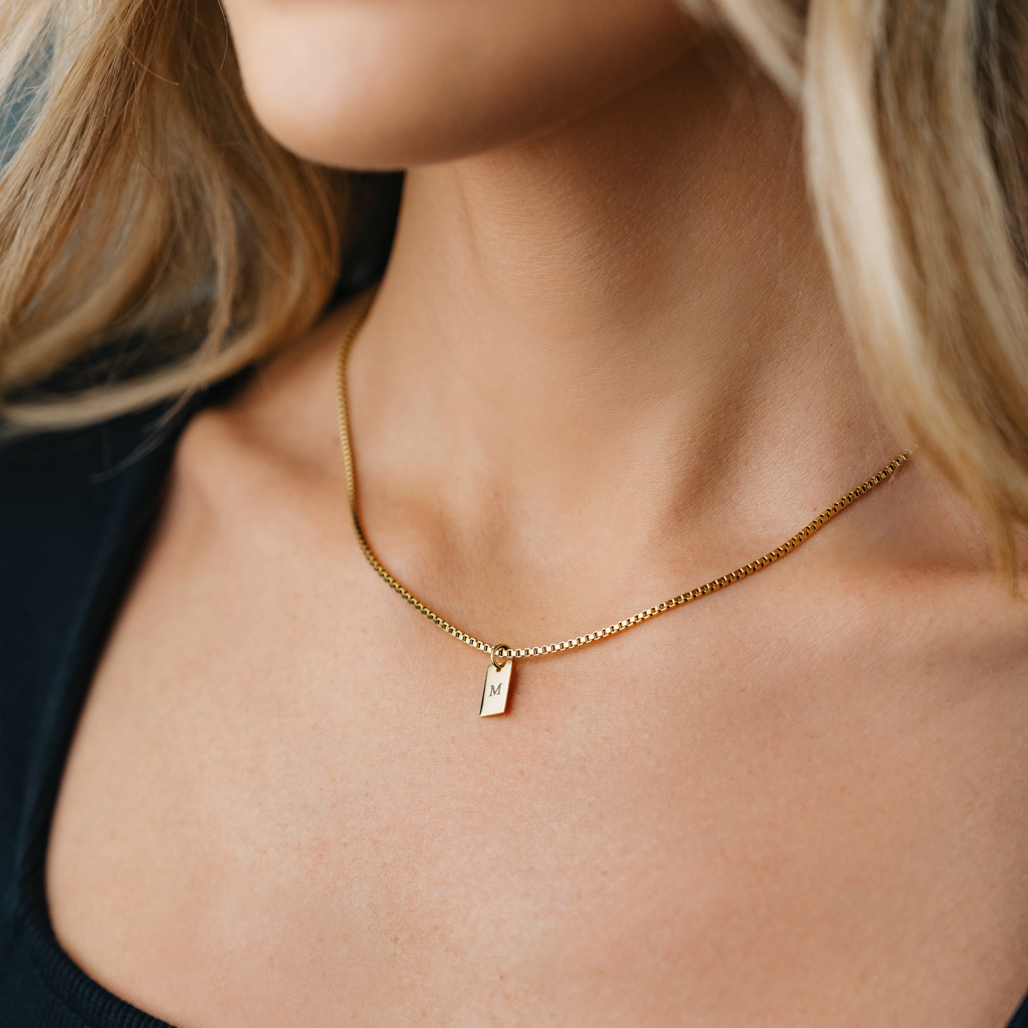 A female wearing SAAR Co's Emily, an 18k Gold Coated, box chain with polished pendant. The pendant has been engraved with the letter M. 
